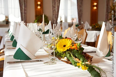 Das Restaurant ist mit langen Tischreihen bestückt. Die Tische sind festlich mit weißer Tischdecke und Servietten mit grün-weiß, sowie Gläsern gedeckt. Am Tischanfang steht eine schöne Blumendeko.