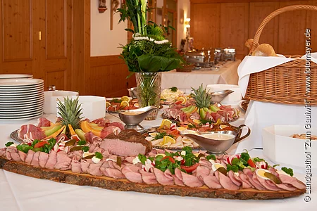 Es leckeres kaltes Buffet ist vor einem Fenster aufgebaut. Es gibt Platten mit Wurst, Lachs und Käse. Erhöht steht der große gut gefüllte Brotkorb. Eine Blumendeko und Teller gehören ebenfalls dazu.