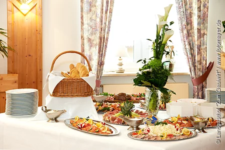 Es leckeres kaltes Buffet ist vor einem Fenster aufgebaut. Es gibt Platten mit Wurst, Lachs und Käse. Erhöht steht der große gut gefüllte Brotkorb. Eine Blumendeko und Teller gehören ebenfalls dazu.