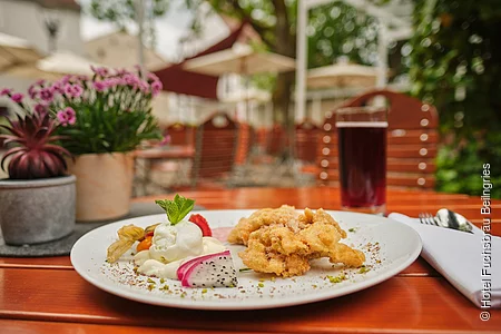 Auf einem Holztisch auf der Terrasse ist eine köstlich angerichtete Portion Obatzda serviert. Im Hintergrund stehen links zwei kleine Blumentöpfe und rechts ein Getränk.