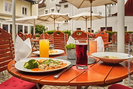 Die Terrasse ist mit Holzmöbeln bestückt. Auf dem vordersten Tisch sind zwei Essen und zwei Getränke angerichtet. Im Hintergrund sieht man die weißen Sonnenschirme.