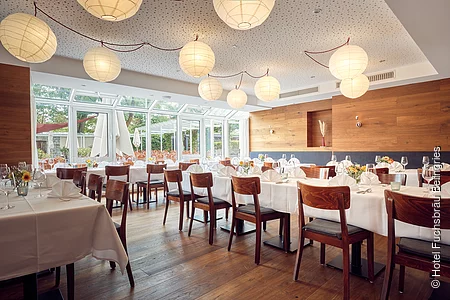 Das Restaurant mit runden, herabhängenden Lampen ist hell und modern eingerichtet. Der dunkle Holzboden passt perfekt zu den Tischen, die mit weißen Tischdecken gedeckt werden. Alle Tische sind festlich gedeckt.