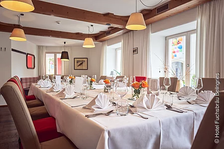 Der Raum mit den herabhängenden Leuchten ist mit einer großen Tafel und braunroten bequemen Stühlen gefüllt. Der Tisch ist festlich mit Tischdecke, weißen Servietten, Geschirr und Weingläsern gedeckt.