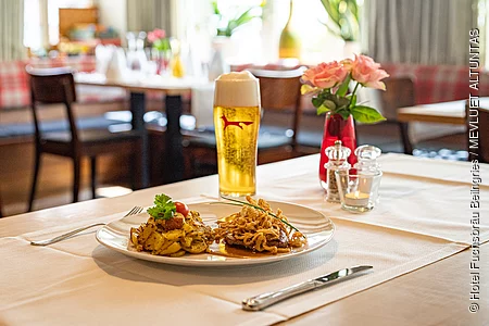 Am Tisch mit beigem Läufer steht ein Helles und auf dem Teller davor ist ein Zwiebelrostbraten mit Bratkartoffeln angerichtet. Im Hintergrund sind weitere Tische zu erahnen.