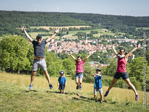 Eine vierköpfige Familie springt mit ausgestreckten Armen und Beinen oberhalb von Berching in die Höhe. Sie landen sanft auf einer grünen Wiese.