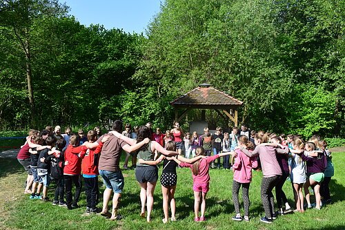 Kinder und Gruppenleiter in einem Kreis, halten sich an den Armen, auf einer Wiese vor Bäumen