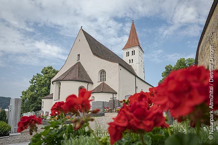 Die Basilika von außen. Rechts ist die Stadtmauer angedeutet und im Vordergrund blühen rote Blumen.