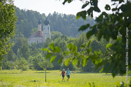 Ein Pärchen wandert unterhalb der Wallfahrtskirche Breitenbrunn entlang satter grüner Wiesen. Die Wallfahrtskirche ist von grünen Wäldern umgeben.