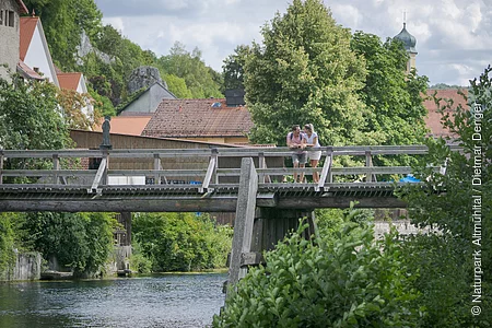 An der Balustrade der Holzbrücke vor Essing lehnt sich ein Pärchen an und schaut flussaufwärts. Hinter  ihnen sieht man einige Hausdächervon Essing umrahmt von Bäumen und deren Blätterdach.