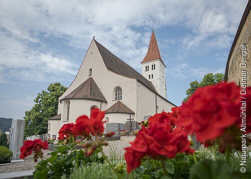 Die Basilika von außen. Rechts ist die Stadtmauer angedeutet und im Vordergrund blühen rote Blumen.