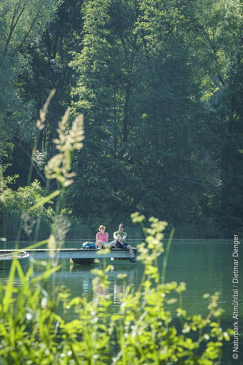 Ein Wanderpärchen sitzt auf dem Steg am Kratzmühlsee und lässt den Blick über den See schweifen. Hinter ihnen ragen die Bäume mit ihrem grünen Blätterkleid auf und links am Steg ist ein Ruderboot befestigt.