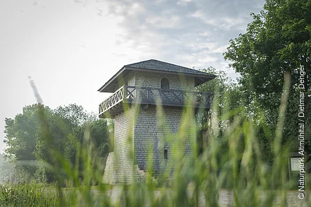 Der Limesturm ist aus weißem Stein rekonstruiert. Die Balustrade ist aus Holz und führt einmal um den ganzen Turm. Es ist unten ein kleines Fenster und oben eine Tür, die auf die Balustrade führt zu sehen. Umrahmt wird der Turm vom grünen Gras und den Bäumen im Hintergrund. Der blaue Himmel ist bewölkt.