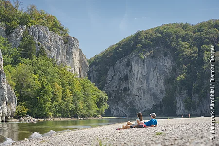 Ein Pärchen liegt am Kiesstrand und lässt seinen Blick zum Donaudurchbruch schweifen. Die Donau fließt zwischen rund 80 m hohen Felswänden hindurch.