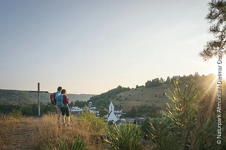 Zwei Wandernde stehen neben dem Kreuz auf dem Pfünzer Kirchberg und schauen über Pfünz zum Osterberg mit seiner Wacholderheide. Über dem Osterberg geht gerade die Sonne unter.