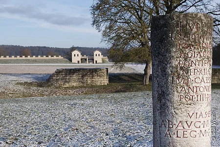 Im Vordergrund befindet sich ein römischer Meilenstein mit Entfernungsangaben zu den nächsten wichtigen Zielen. Im Hintergrund der winterlichen Landschaft ist die rekonstruierte Toranlage des römischen Kastells Vetoniana zu sehen.