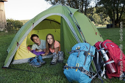Eine Frau und ein Mann liegen im Zelt und schauen aus diesem heraus. Vor dem Zelt liegen ihre zwei Wanderrücksäcke.