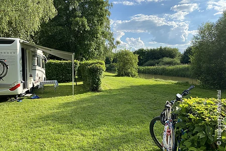 Auf dem Campinplatz steht links ein Wohnmobil mit  herausgelassener Jalousie. Hinter dem Mobil ragen große Bäume auf. Die Wiesen und Sträucher strahlen im hellen grün. Rechts lehnt ein Fahrrad an einem Strauch.