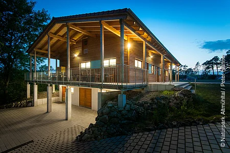 Ein Holzhaus mit großer Terrasse ringsherum ist hellbeleuchtet in der Nacht. Ein gepflasteter Weg führt links hinunter zum selben Gebäude, das im Untergeschoss gemauert und im Hang gebaut ist. Die Terrasse wird durch Pfeiler gestützt.