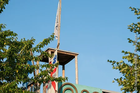 Eingerahmt von zwei großen Bäumen steht ein Aussichtsturm mit einem Kunstobjekt auf grünem Hintergrund. Links ist eine Fahne gehisst.