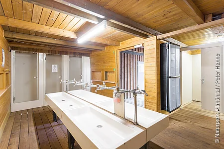 Der Waschraum hat zwei große Waschbecken in der Mitte . Rechts geht es zu den Duschen und WC's. Das Gebäude ist aus Holz gebaut.