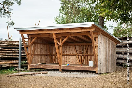Eine Schutzhütte aus Holz steht vor einem Holzzaun. Dahinter stehen einige Bäume.