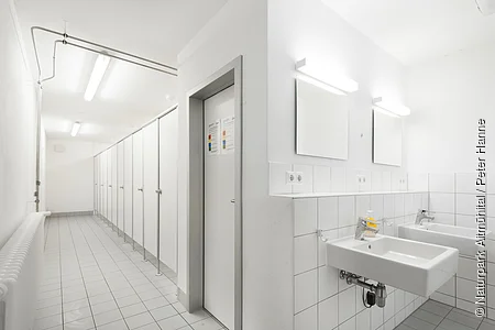 Der Waschraum ist komplett in weiß gehalten. Links ist ein langer Gang mit lauter Türen, die rechts zu Umkleiden und WC's führen.