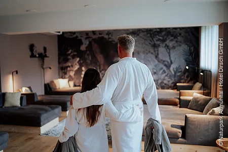 Eine Frau und ein Mann im Bademantel mit Handtücher im Ruheraum des Hotel Dirsch in Emsing. Im Hintergrund Liegen und ein Sofa mit einer kleinen Beleuchtung im Raum.