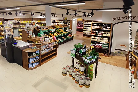 Das Innere des Verkaufsraumes des Hofladens. In der Mitte die Kasse umgeben von Holzregalen. Die Waren sind schön aufgeräumt in weiteren Holzregalen. Ganz rechts befindet sich die Gemüsetheke
