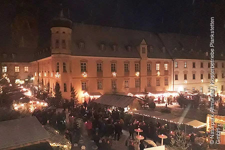 Kloster Plankstetten bei Nacht Vor dem Gebäude sind Stände Adventsstände aufgebaut, belebt mit Menschen. Alles ist weihnachtlich beleuchtet.