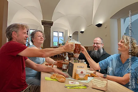Vier Personen, zwei Frauen und zwei Männer stehen um eine Holztheke, und prosten sich mit einem Krug Bier zu. Auf der Theke stehen eine Kiste mit Besteck und ein kleiner Korb. Es liegen zwei Semmeln und eine Breze daneben. Im Hintergrund sieht man Säulen und Bügen des Klostegewölbes.