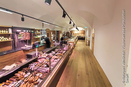 Eine Fleisch- und Wursttheke mit Inhalt seitlich aufgenommen. Dahinter ein Holzregal mit verschiedenen Waren.