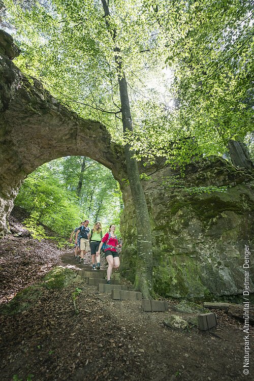 Eine kleine Wandergruppe steigt die Stufen durch das Felsentor hinunter. Hinter dem Tor erstreckt sich der Wald, wo die Sonne ein wenig durchblitzt.