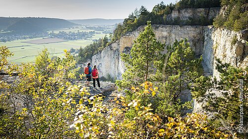 Zwei Leute mit Rucksack stehen zwischen Bäumen nahe der Abbruchkante des ehemaligen Steinbruchs und blicken auf die Wand. Im Hintergrund breitet sich das Altmühltal aus.