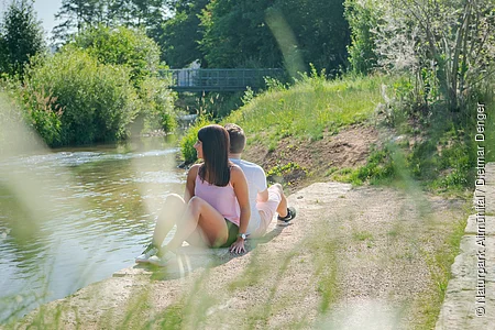 Ein junges Paar sitzt entspannt am Ufer der Altmühl, mit dem Rücken zur Kamera. Die Frau trägt ein rosa Oberteil, der Mann ein weißes T-Shirt. Beide blicken in die grüne Flusslandschaft, die von Bäumen und Sträuchern gesäumt ist. Im Hintergrund führt eine kleine Brücke über das Wasser. Die Szene ist von hohem Gras im Vordergrund leicht verschwommen eingerahmt.