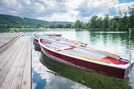 Rote Boote liegen am Holzsteg am Kratzmühlsee. Zwei Wanderer liegen am Steg, der Himmel spiegelt sich im ruhigen Wasser. Am rechten Ufer stehen Bäume.