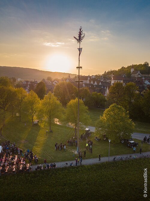 Auf dem Bild sieht man das Aufstellen des Maibaums in Greding. Im Hintergrund sieht einen Sonnenuntergang.