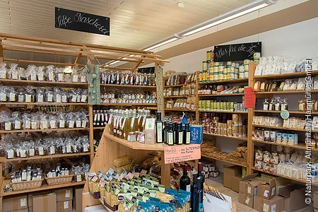 Blick in den Hofladen. Hohe Holzregale, gefüllt mit regionalen Produkten in Tüten, Dosen und Gläsern. Davor ein Tisch, auf dem weitere eingepackte Produkte präsentiert werden.