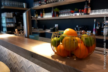 Die Bar ist aus Holz. Die Regale sind gut gefüllt mit leckeren Spirituosen und Gläsern. Auf dem Tresen steht ein Obstkorb als Dekoration.
