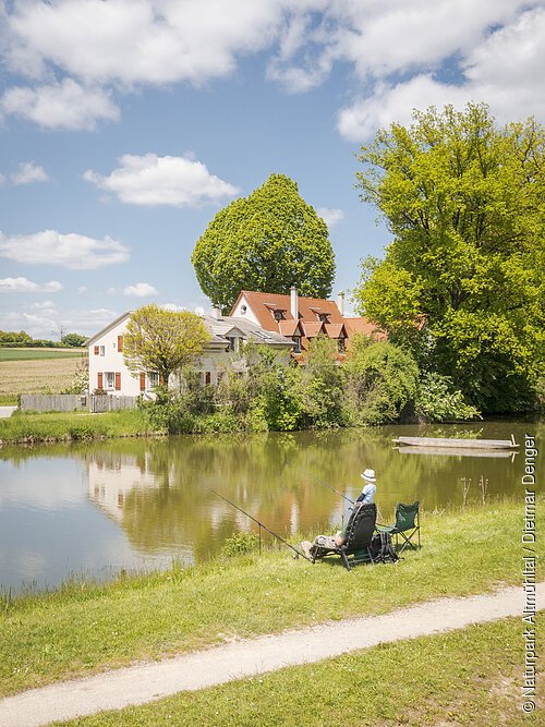 Ein Angler mit seiner Angel sitzt am Ufer der Altmühl und wartet darauf, dass ein Fisch anbeißt.