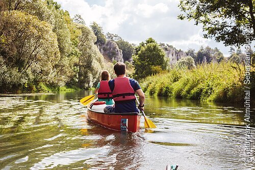Ein Mann und eine Frau mit dem Rücken zu uns, sitzen in einem roten Kanu auf der Altmühl. Die gelben Paddel erzeugen leichte Wellen auf der Wasseroberfläche. im Hintergrund ist das grüne Ufer mit prächtigen Bäumen zu sehen und rechts blitzt der blaue Himmel durch.