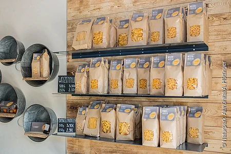Rechts sind drei Regalbretter an einer Holzwand angebracht. Darauf stehen mehrere Nudelpackungen. Daneben an einer weißen Wand sind verschieden große Zinswannen mit Innenbrett als Regale angebracht, in denen sich Regionale Produkte wie Eierlikör, Eier und Nudelpackungen befinden.