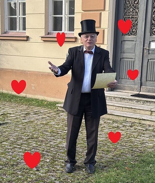Valentinstagsführung 2026 Ein Mann in Frack und Zylinger liest aus einem vermeintlichen Liebesbrief vor. Er ist von roten Herzen umgeben ...
