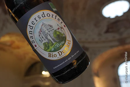 Flasche Sandersdorfer Bio Dunkel Bier mit Etikett, im Hintergrund unscharfe Architektur.