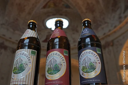 Drei Bierflaschen mit Etiketten der Marke Sandersdorfer, im Hintergrund eine verzierte Decke.