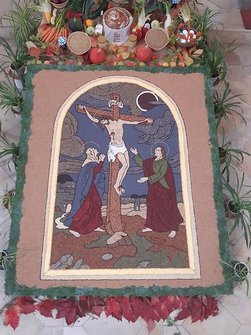 Der Körnerteppich zeigt Jesus am Kreuz mit seiner Mutter Maria und dem Jünger Johannes.