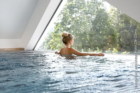 Schwimmbad Hotel Die Gams Ein Gast genießt den Ausblick von Schwimmbecken durch ein Fenster nach draußen in die Naturlandschaft.