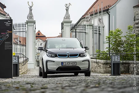 Ein Elektroauto steht auf einem gepflasterten Weg vor einem historischen Gebäude mit Statuen und einem Tor.