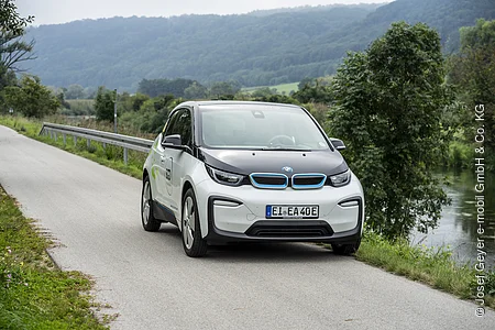 Ein weißes Elektroauto fährt auf einer ländlichen Straße entlang eines Flusses, umgeben von Bäumen und Hügeln.