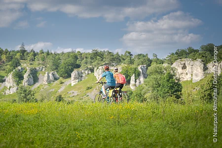 Zwei Personen fahren mit Fahrrädern auf einem grasbewachsenen Feld vor einer Felslandschaft.