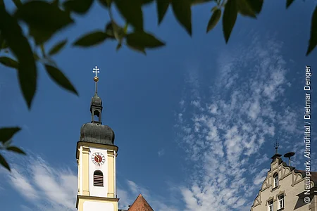Heilig-Kreuz-Kirche in Altmannstein Kirche mit Turm und Uhr, umgeben von Gebäuden und Bäumen, unter blauem Himmel mit Wolken.